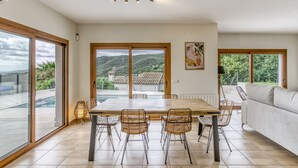 A spacious dining area with a large table and chairs, large windows with a view of hills, and a white sofa. - Villa 'Maréa' with Sea Views, Wi-Fi, and Air Conditioning (Calonge)