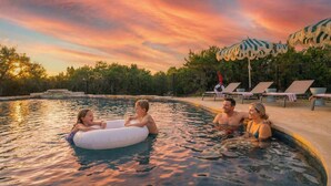 Piscina con sillas de descanso y sombrillas, rodeada de árboles y un cielo al atardecer.
