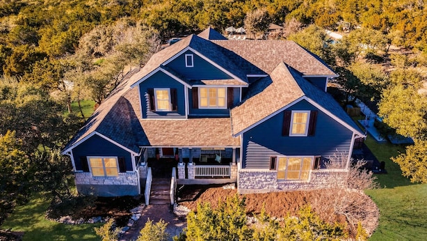 Una casa de dos pisos con base de piedra y revestimiento azul, rodeada de un área boscosa.