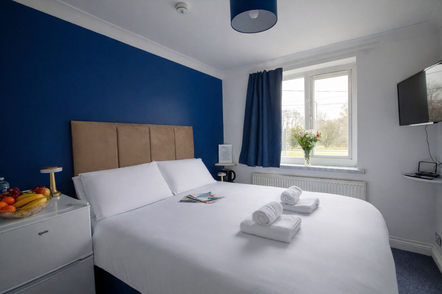 Une chambre d’hôtel avec un lit, une table de chevet avec des fruits, une lampe, une fenêtre avec des rideaux et un téléviseur.