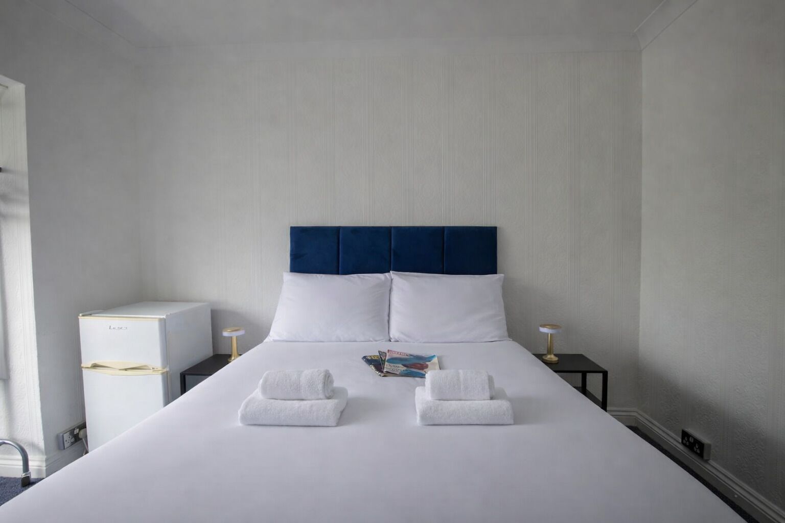 A neatly made bed with a blue headboard, white pillows, and two folded towels on the bed. There is a small bedside table with a lamp and a box on the floor beside the bed.