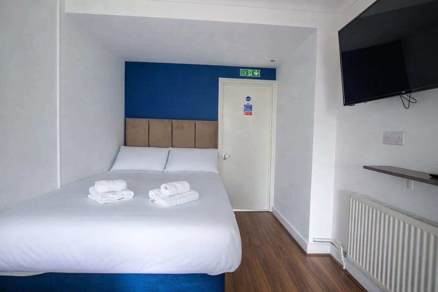 A hotel room with a bed, a TV mounted on the wall, and a door with an emergency exit sign.