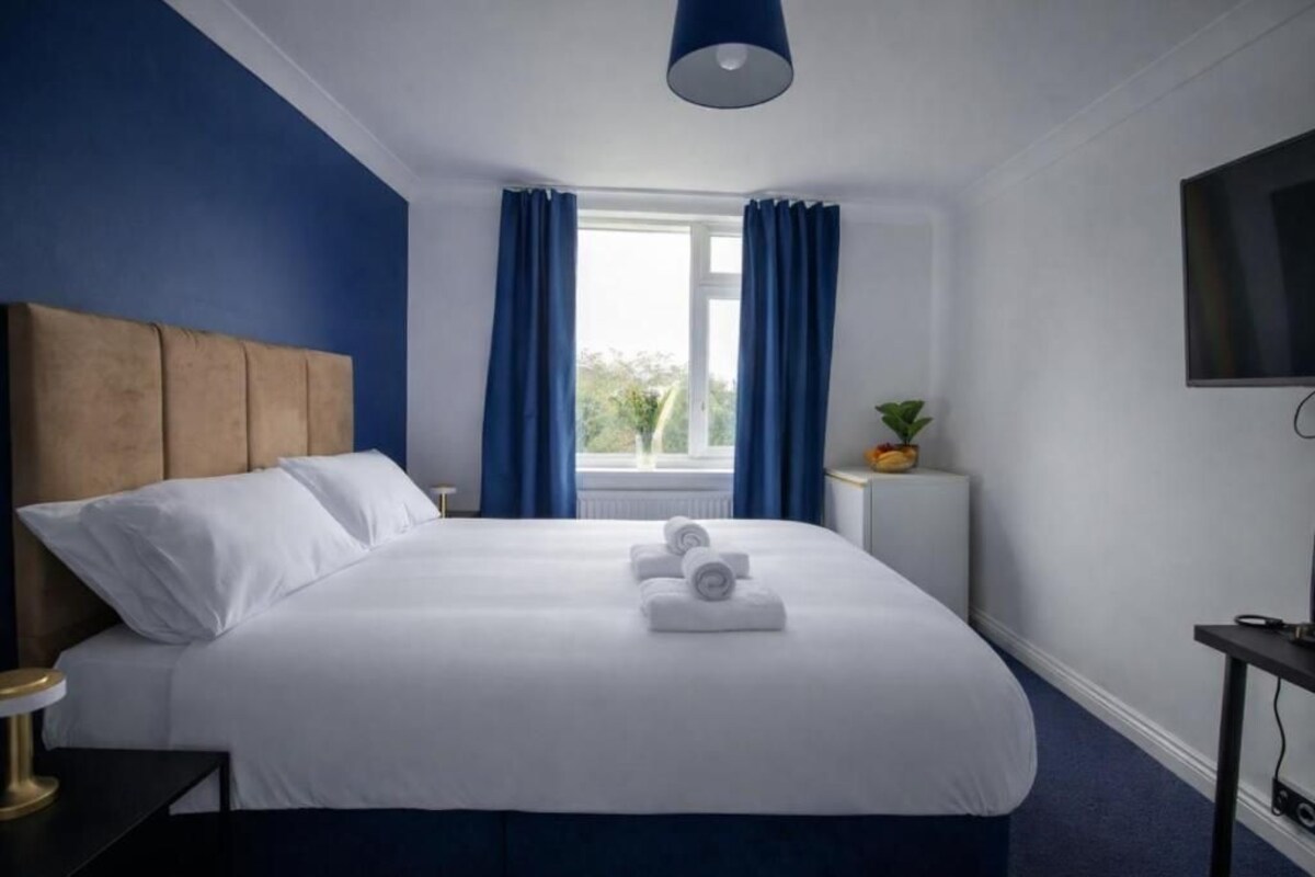 A hotel room with a large bed, a headboard, a bedside table with a lamp, a small table with a plant, and a TV on the wall.