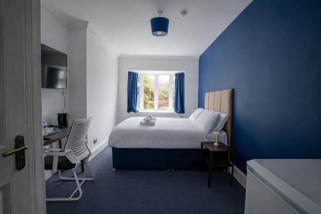 A hotel room with a bed, a desk, a chair, a TV, a window with curtains, and a blue accent wall.