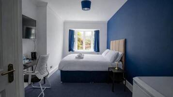 A hotel room with a bed, a desk, a chair, a TV, a window with curtains, and a blue accent wall.