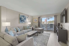Ein Wohnzimmer mit einer Sofagarnitur, einem Couchtisch, einem Fernseher und einem Balkon mit Blick auf einen Leuchtturm.