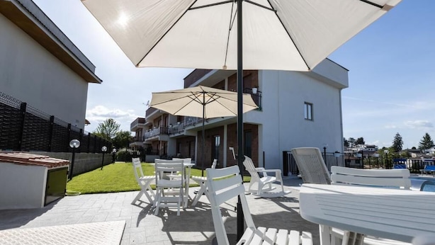An outdoor patio with white lounge chairs and a large umbrella, surrounded by a swimming pool and residential buildings.