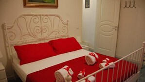 Una cama bien hecha con una colcha roja y almohadas blancas, un cabecero de metal blanco y toallas enrolladas con cintas rojas sobre la cama.