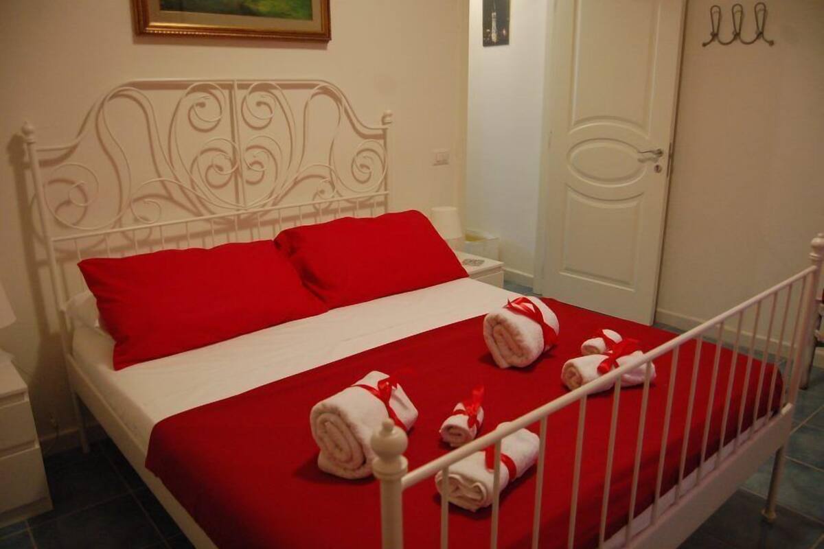 Una cama bien hecha con una colcha roja y almohadas blancas, un cabecero de metal blanco y toallas enrolladas con cintas rojas sobre la cama.