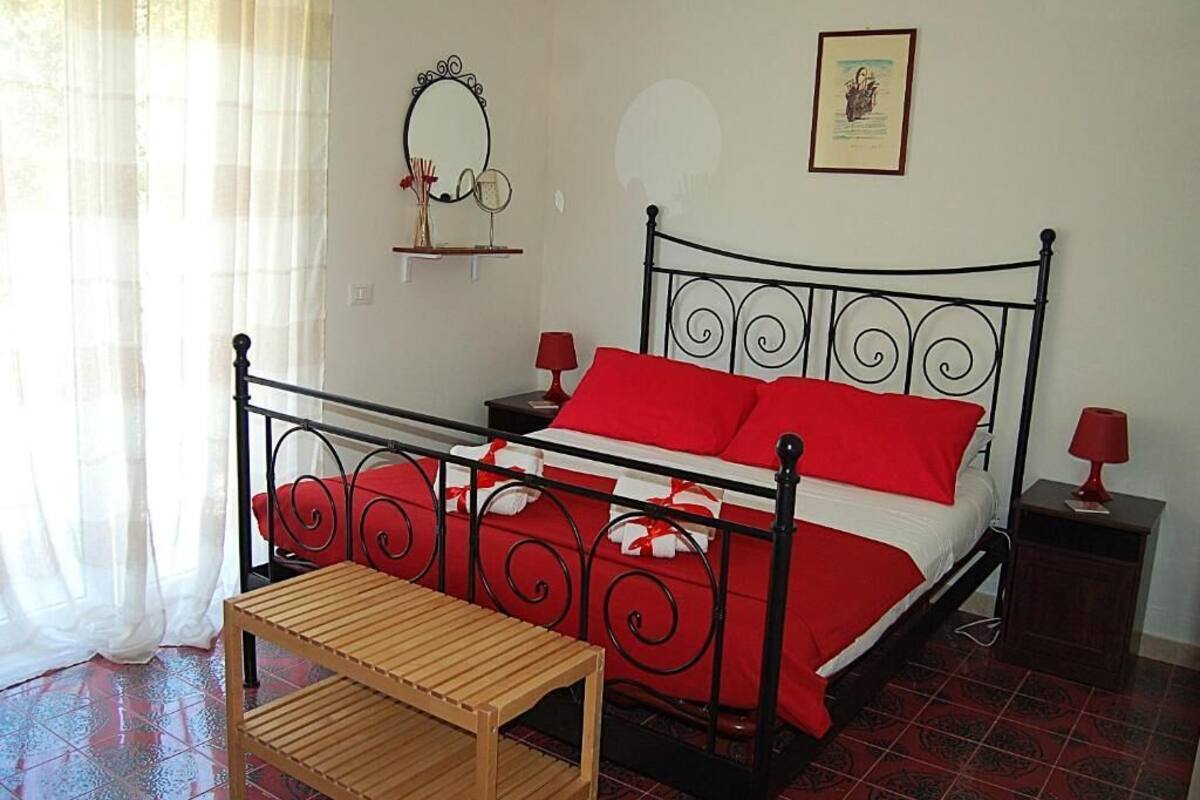 Un dormitorio con estructura de cama de metal, ropa de cama roja, una mesa de madera y una mesita de noche con lámpara.