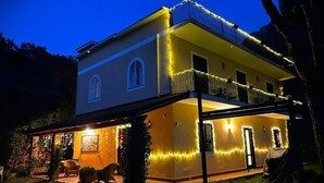 Una casa de dos pisos con balcón, iluminada por luces colgantes, y un espacio de estar al aire libre cubierto.