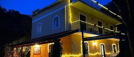Uma casa de dois andares com varanda, iluminada por luzes de fada, e uma área de estar coberta ao ar livre.