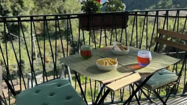 Un balcón con una mesa lista para dos personas, que incluye un recipiente con aperitivos, una copa de bebida roja y una pequeña planta.