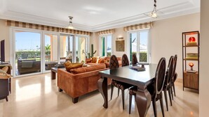 A spacious living area with a large sofa, dining table, and chairs. There are sliding glass doors leading to a balcony with a view of the sea. - Sotogrande Marina Duo Apartment (Sotogrande)