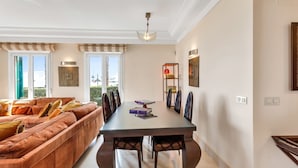 A modern living room with a sofa, dining table, and chairs. There is a window with blinds, a ceiling light, and a wall-mounted shelf with decorative items. - Sotogrande Marina Duo Apartment (Sotogrande)