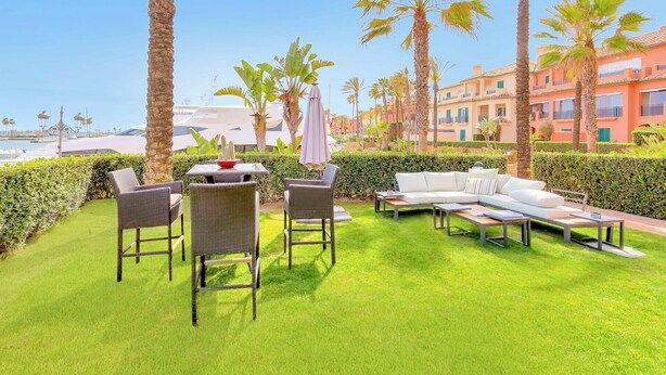 An outdoor seating area with wicker chairs and a white sofa, surrounded by palm trees and a well-maintained hedge. - Sotogrande Marina Duo Apartment (Sotogrande)