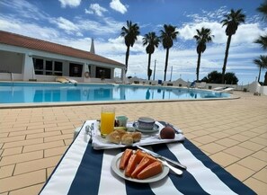 A poolside breakfast setup with a tray of food, a glass of orange juice, and a view of the pool and palm trees. - Hotel Albergaria Dom Manuel (Lagoa)