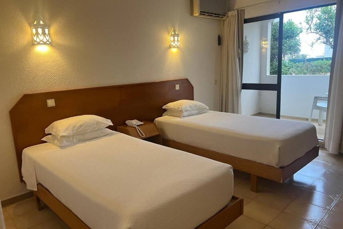 Una camera d'hotel con due letti singoli, una testiera in legno, un telefono sul comodino e vista all'esterno da una grande finestra.