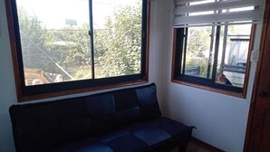 A room with a dark blue sofa, two windows with wooden frames, and a view of trees and a construction site outside. - Lomas del Hualle - House 2 (Valdivia)