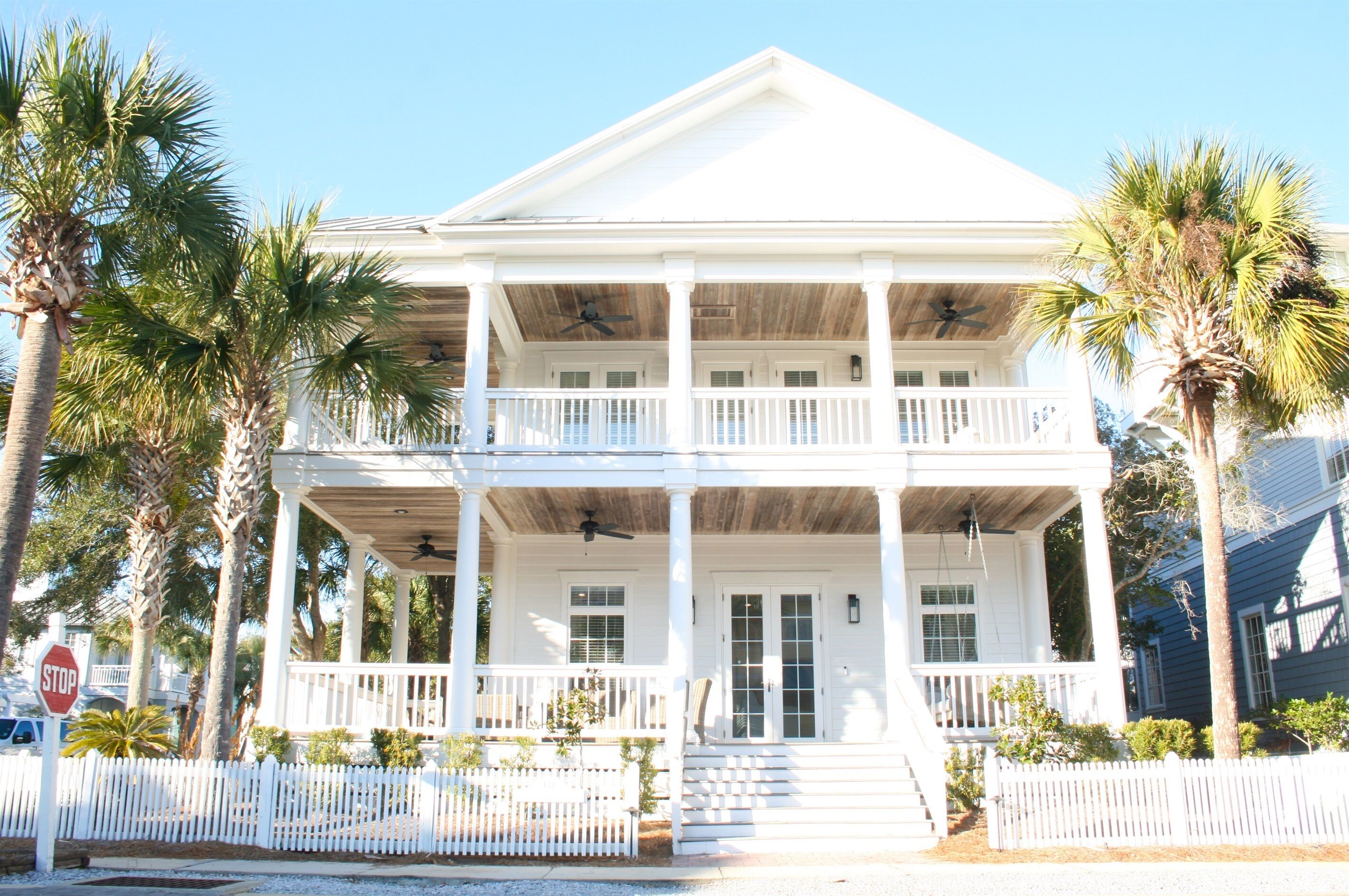 A two-story white house with a balcony, palm trees, and a white picket fence.