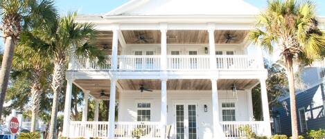 A two-story white house with a balcony, palm trees, and a white picket fence.
