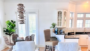 A modern living room with two light grey sofas, a round coffee table, wicker chairs, a hanging light fixture, and a kitchen area with white cabinets and a black countertop. - Charming Carillon Home + Kid Friendly + Lots of Extras (Panama City Beach)