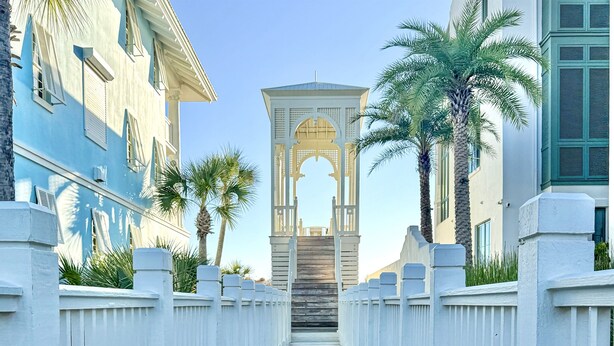 A pathway leading to a central archway flanked by palm trees and buildings with white and blue facades. - Charming Carillon Home + Kid Friendly + Lots of Extras (Panama City Beach)