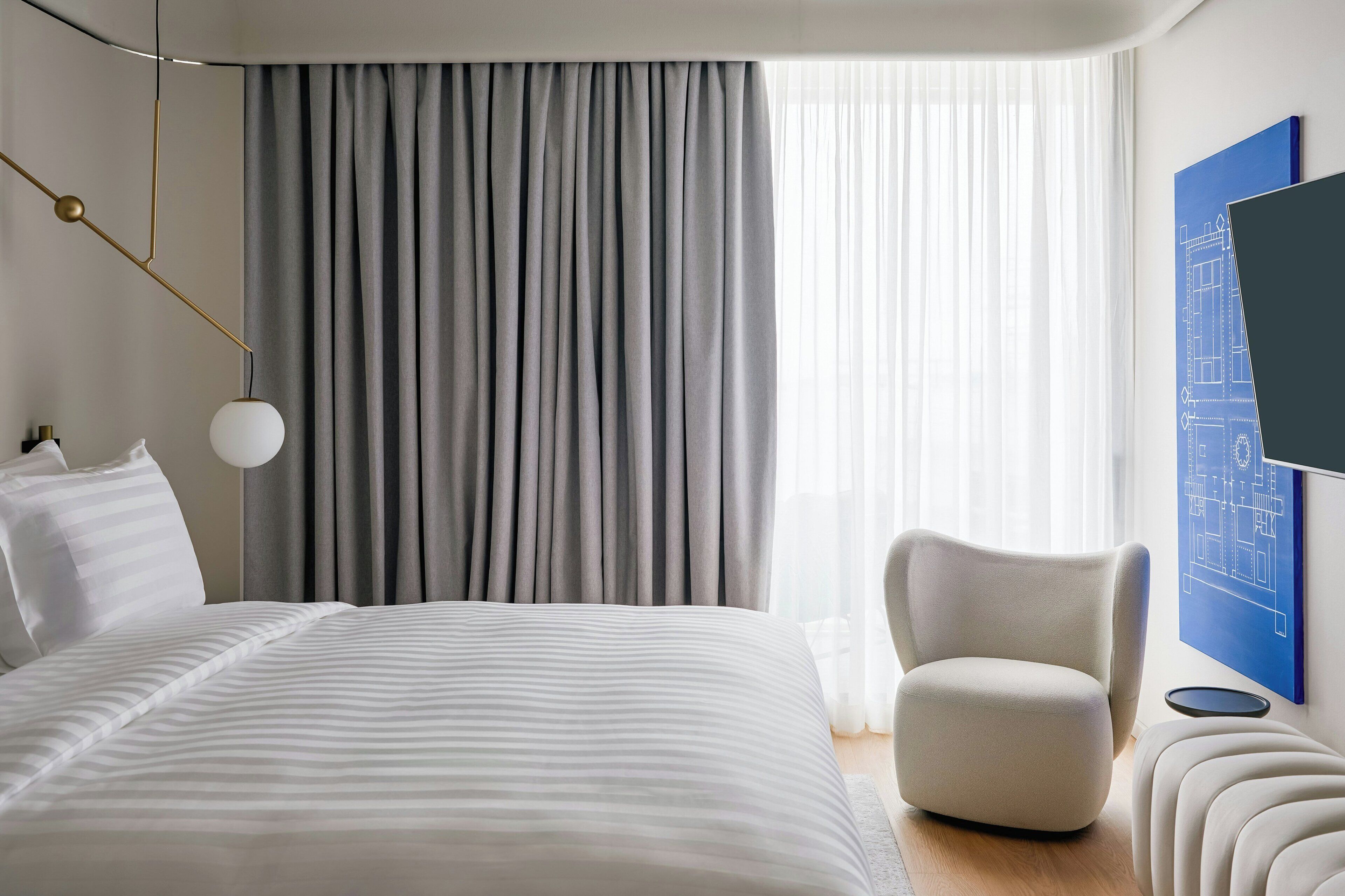 A modern hotel room with a large bed, a chair, a desk, and a large window with curtains.