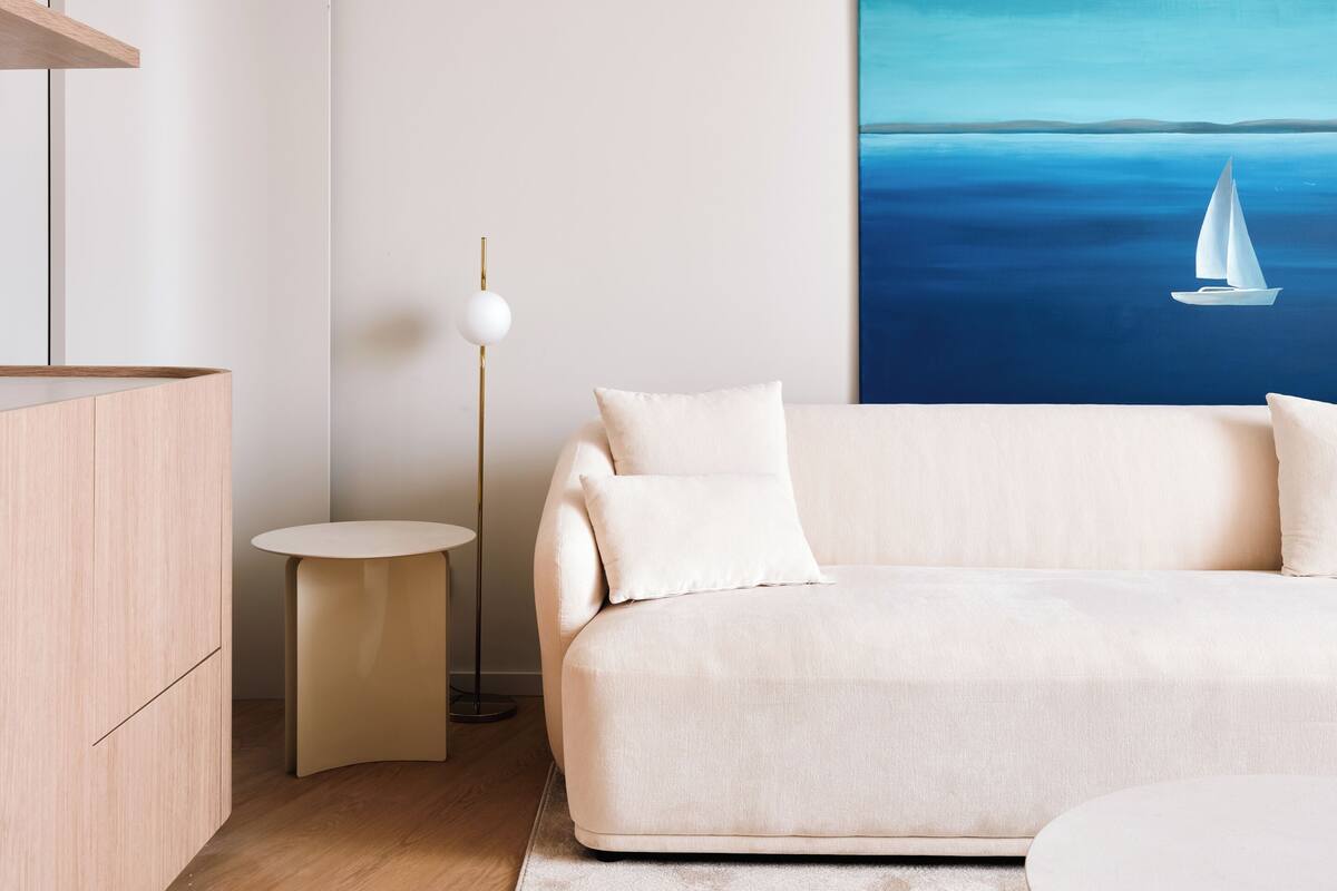 Una sala de estar moderna con un sofá, una mesa redonda de centro, una mesita auxiliar, una lámpara de pie y un cuadro de un velero en el mar.