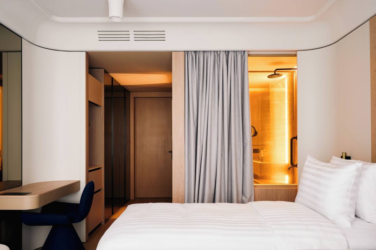 Habitación de hotel con cama, escritorio, silla, zona de ducha y armario.