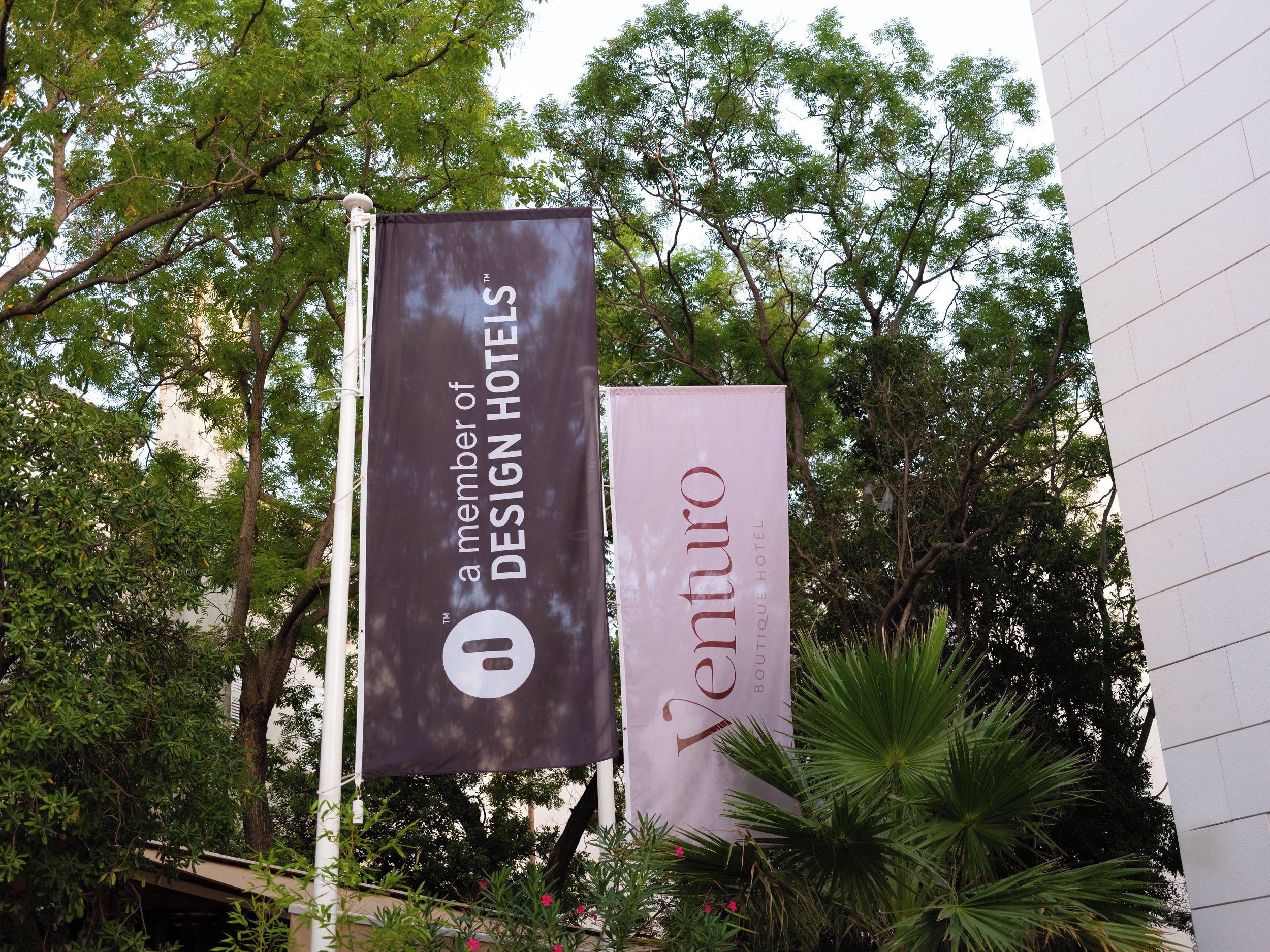 Zwei Banner für 'The S' und 'Venturos Boutique Hotel' mit umgebender Bepflanzung.