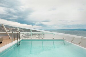 En swimmingpool med en metalstige, en hvid terrasse og udsigt til havet og fjerne kyster.