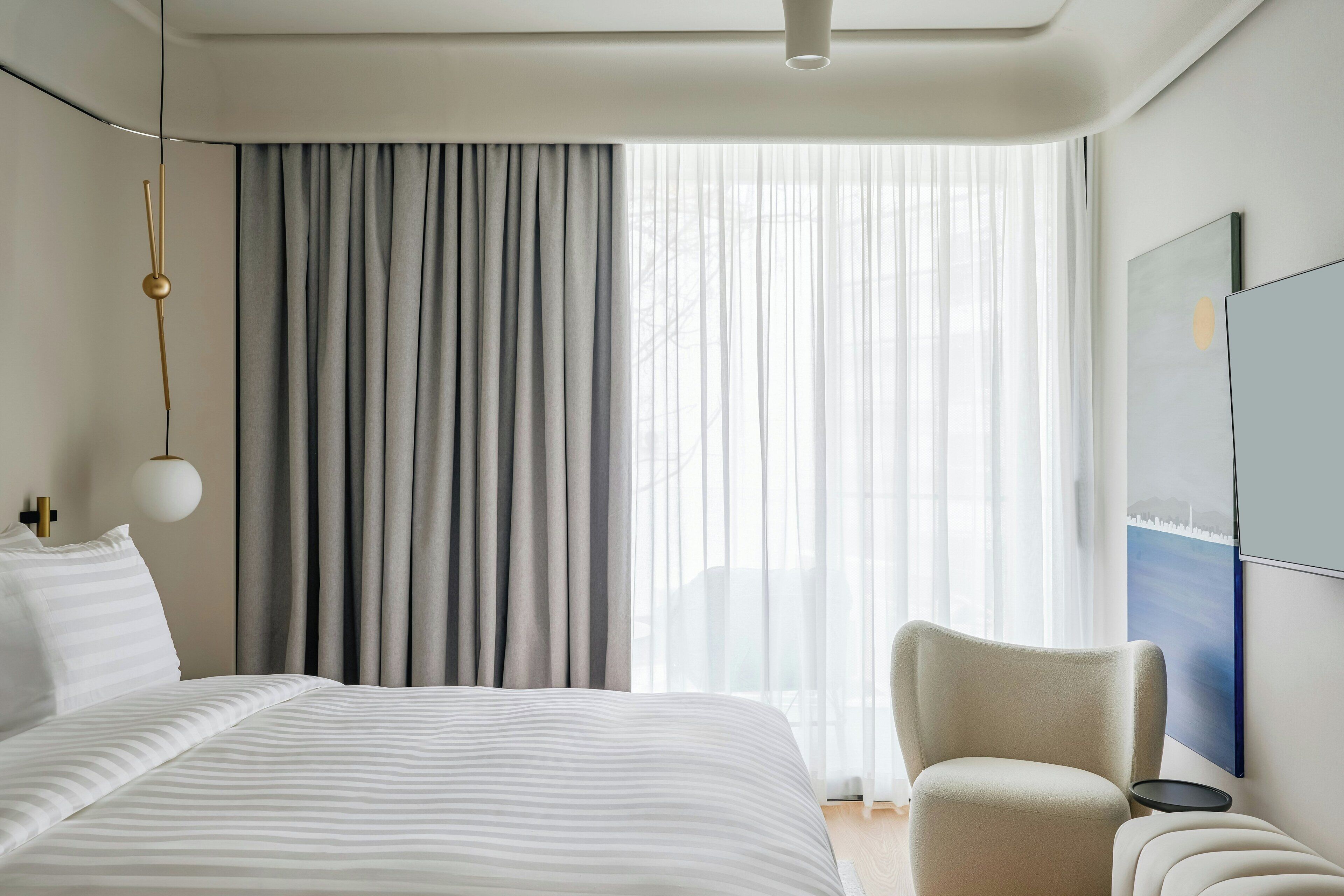 A modern hotel room with a large bed, a chair, a small table, and a painting on the wall.