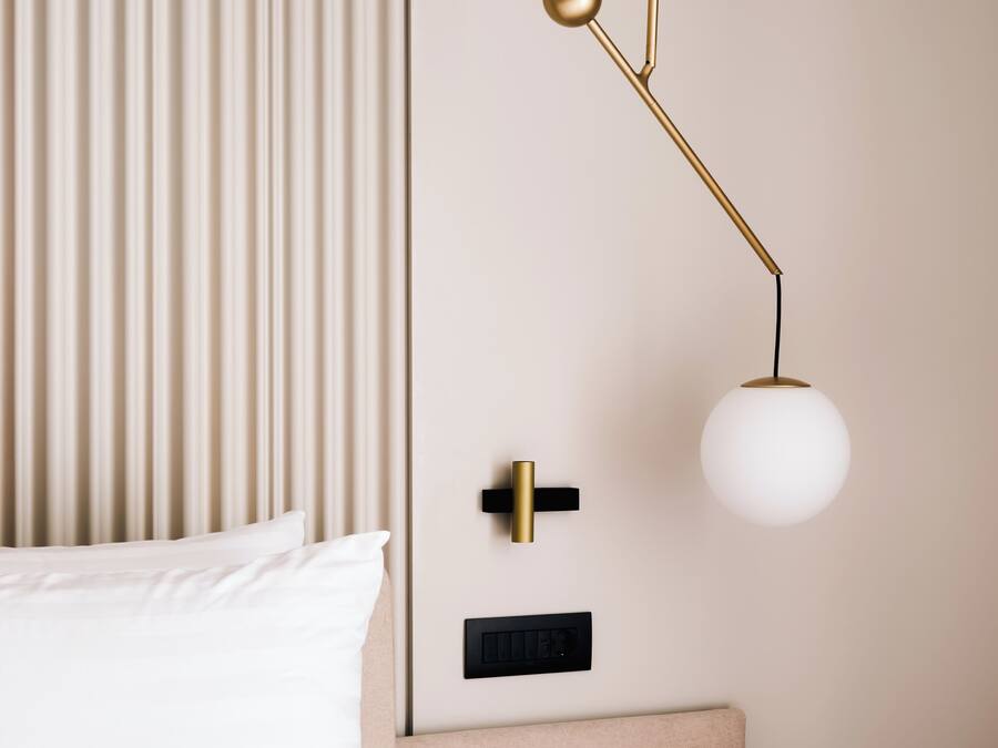 A bed with white linens, a bedside table with a lamp, and a wall-mounted control panel.