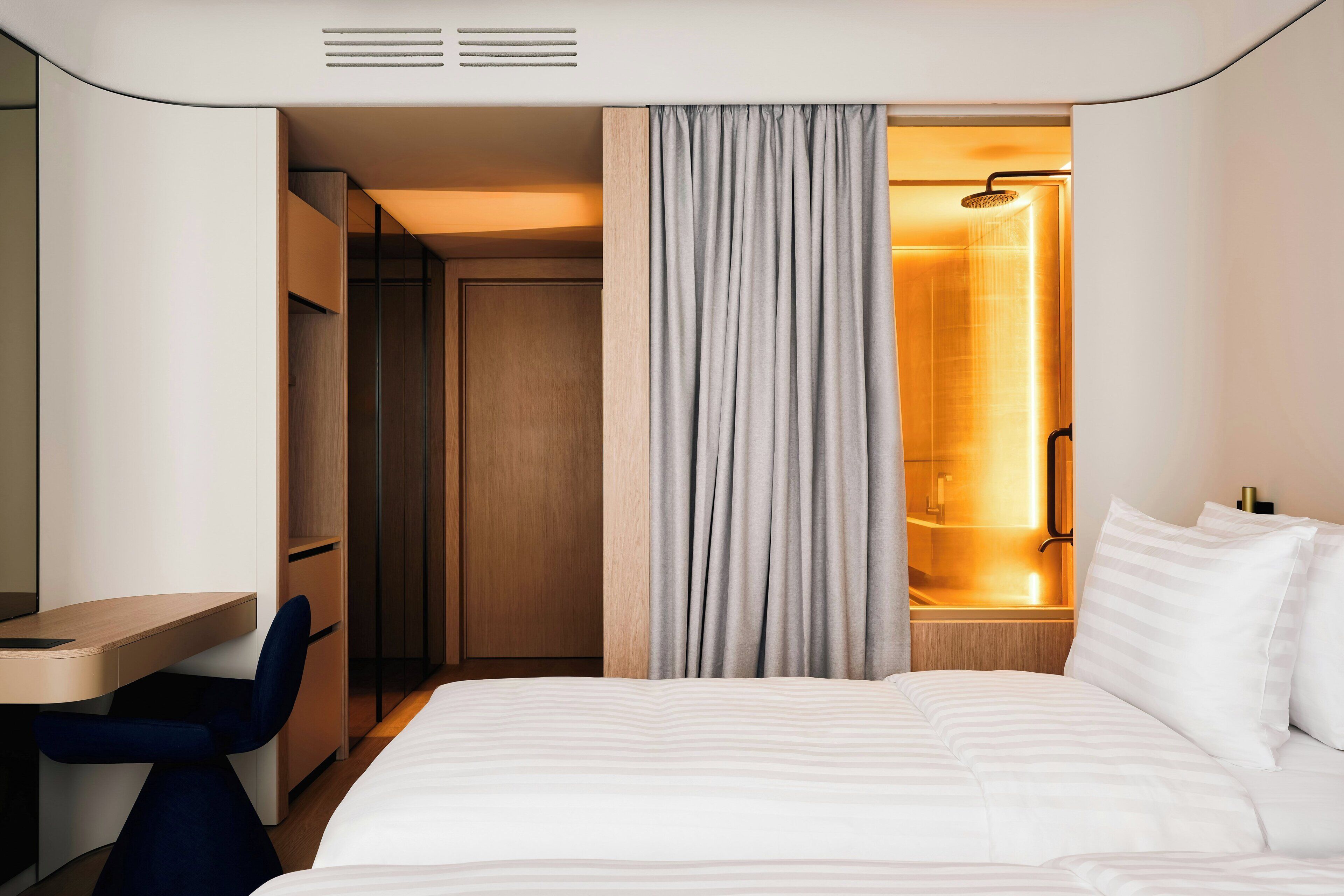 A hotel room with a bed, a desk, a chair, a wardrobe, and a shower area.
