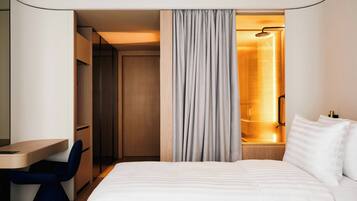 A hotel room with a bed, a desk, a chair, a wardrobe, and a shower area.