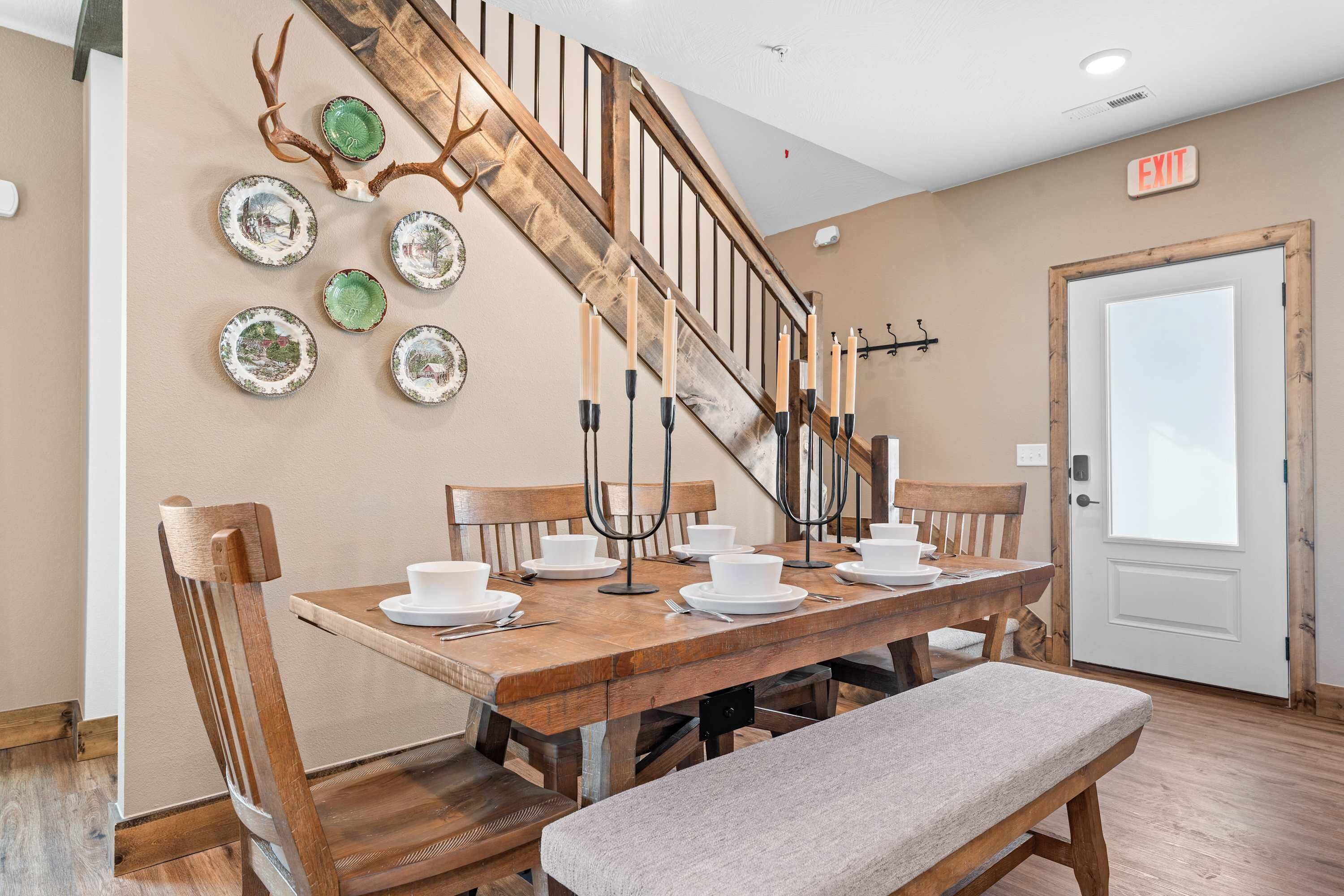 A dining area with a wooden table set for four, surrounded by chairs. A bench is placed in front of the table. A staircase with a wooden railing and antlers decor is visible in the background.