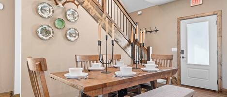 A dining area with a wooden table set for four, surrounded by chairs. A bench is placed in front of the table. A staircase with a wooden railing and antlers decor is visible in the background.