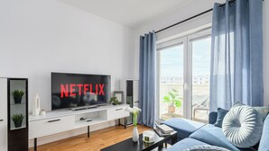 A modern living room with a flat-screen TV displaying "NETFLIX," a sofa, a coffee table, and a large window with curtains. - Lumina 3-rooms apart with balcony (Łódź)