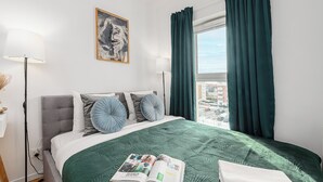 A neatly arranged hotel room with a bed, a window with curtains, a floor lamp, and a painting on the wall. - Lumina 3-rooms apart with balcony (Łódź)