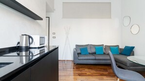 A modern kitchen with a microwave, kettle, and black countertop. There is a grey sofa with blue cushions and a glass table. The room has wooden flooring and white walls. - GuestHost - A cozy and bright apartment of approximately 35 square meters, perfect for two people, located on the second floor of a building with an elevator (NOT accessible to disabled guests).The property is located in a central area of ​​Milan and (Milan)