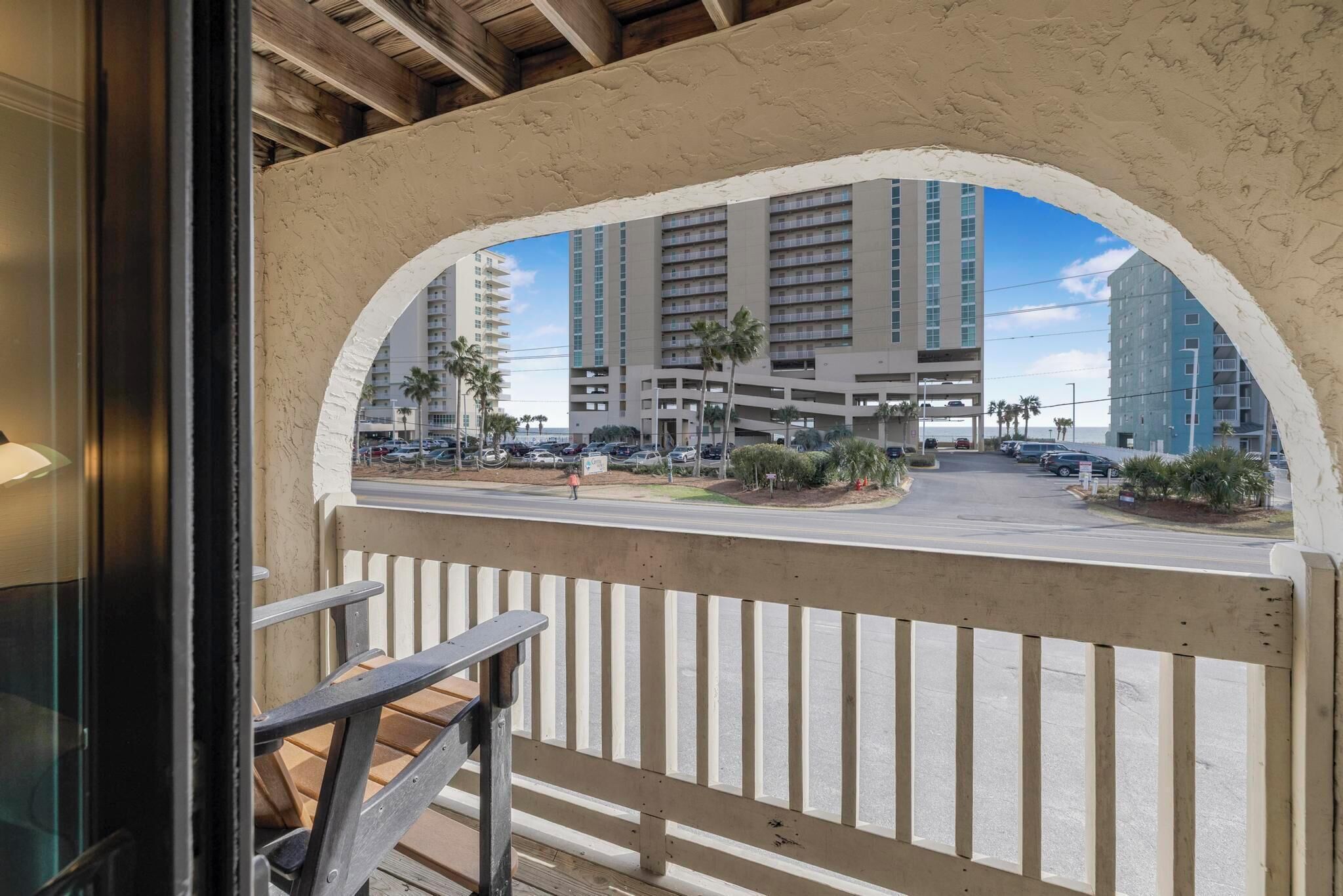 A balcony with a view of a multi-story building, a parking area, and palm trees.