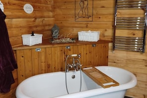 Une salle de bain rustique, avec des murs en bois, une baignoire blanche, un porte-serviettes et une étagère en bois où sont rangés les articles de toilette.