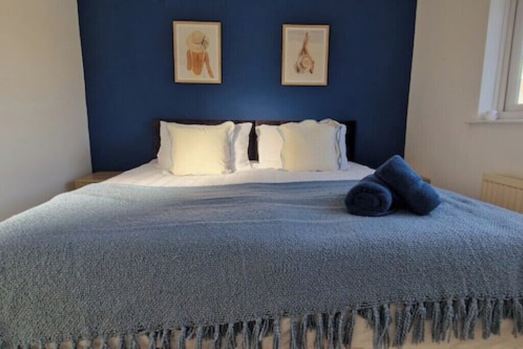 Ein Bett mit blauer Decke und Kissen, zwei gerahmte Bilder an der Wand und ein Fenster zur Rechten.
