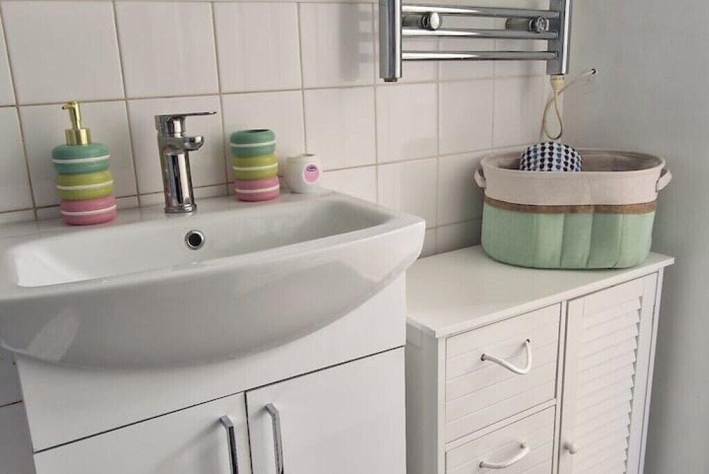 Un baño con un lavamanos blanco, un grifo cromado y un perchero. Hay dispensadores de jabón de colores y un recipiente a rayas en la encimera.