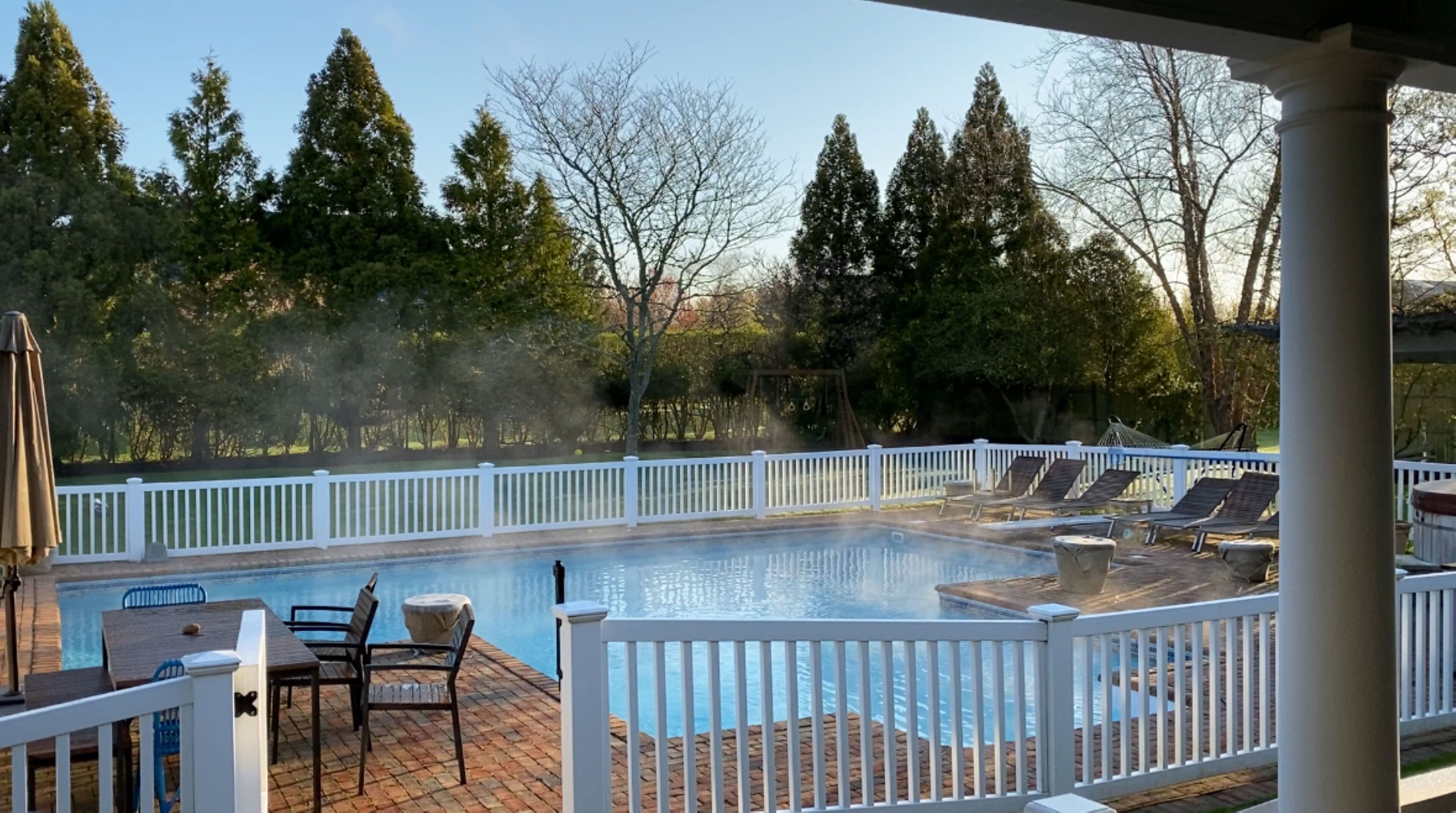 A swimming pool with a white deck and lounge chairs, surrounded by a white fence and trees.