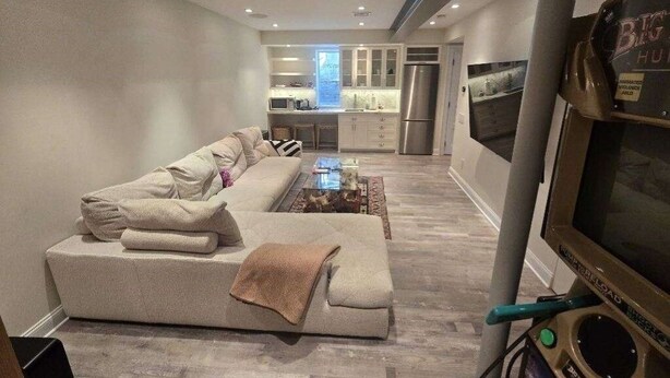 Ein modernes Wohnzimmer mit einer Couch, einem Couchtisch und einem Fernseher. Im Hintergrund ist eine Küche mit Kühlschrank und Mikrowelle zu sehen.