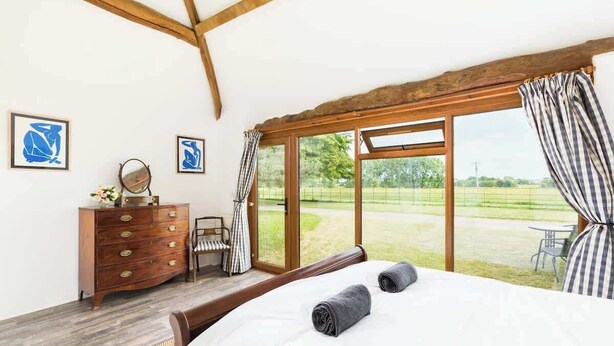 A bedroom with a bed, wooden dresser, and a view of a grassy area through large windows. - Rural, Self-Contained 1Bed, The Writer's Retreat (Hampton Lucy)