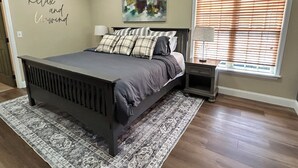 Een slaapkamer met een bed, een nachtkastje, een raam met jaloezieën, een vloerkleed en een muur met een schilderij.