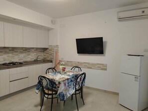 Une cuisine de taille modeste, avec des meubles blancs, un crédence carrelée, une cuisinière à gaz, un évier, une table recouverte d’une nappe à motifs floraux, deux chaises, un téléviseur à écran plat et un réfrigérateur blanc.